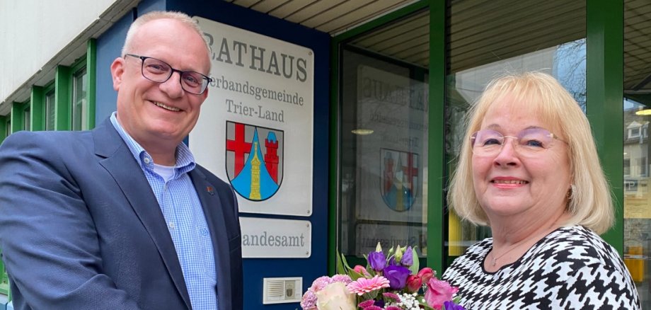 Bürgermeister Michael Holstein überreicht Herta Kartheiser einen Blumenstrauß. Im Hintergrund ist das Wappen der Verbandsgemeinde Trier-Land zu sehen.